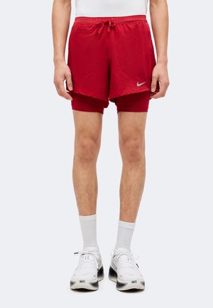 Homme portant un short de course rouge superposé, des chaussettes blanches et des chaussures de course blanches Nike, debout sur un fond clair uni.