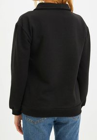 Sudadera negra con un corte relajado, puños y dobladillo de canalé, que exhibe una textura lisa, llevada sobre jeans azules.