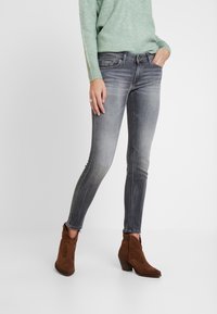 Žena oblečená v vybledlých černých skinny džínách, zeleném pleteném svetru a hnědých semišových kotníkových botách stojí na bílém pozadí.