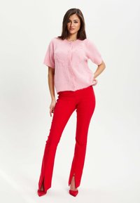 Light pink knitted short-sleeve cardigan with button front, paired with red fitted pants featuring side slits and matching red heels.