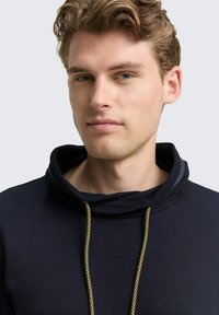 Marineblauwe hoodie van een textuurstof, met een grote kraag en een gouden trekkoord. Close-up van de halslijn en gezichtskenmerken.