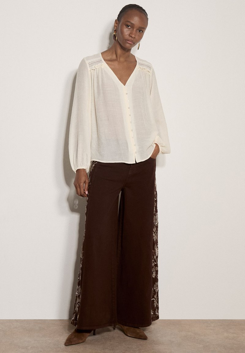 Femme portant une blouse blanche à manches longues et un pantalon marron à jambes larges avec des panneaux latéraux fleuris, debout avec une main dans la poche contre un mur uni.
