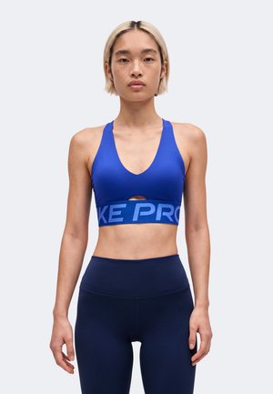 Mujer con el pelo rubio corto, vistiendo un sujetador deportivo azul y leggings azul oscuro, de pie y mirando hacia adelante con expresión neutral.