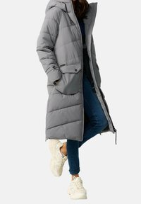 Jasnoszary, ocieplany długi płaszcz z kapturem, z dwoma przednimi kieszeniami i zapięciem na suwak. Noszony na ciemnych dżinsach i jasnych sneakersach.