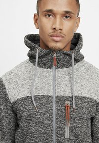 Sudadera con cremallera en gris y negro con un acabado texturizado, bloques de color en contraste y capucha con cordón. Presenta un bolsillo en el pecho con cremallera.
