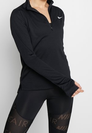 Žena nosící černý polovypínací top Nike s dlouhým rukávem a černé síťované legíny Nike Air, stojící před jednobarevným světlým pozadím.