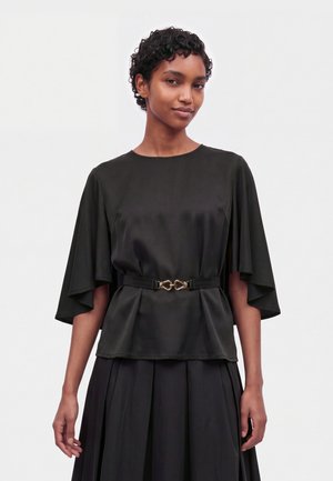 Modèle portant un chemisier noir aux manches fluides façon cape, une ceinture rehaussée de détails dorés et une jupe noire plissée, sur fond uni.