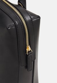 Musta nahkalaukku, jossa on jämäkkä muoto, koristettuna kultaisella metallivetoketjulla ja nahkaisella vetimellä, korostaen siistejä ja yksinkertaisia muotoiluelementtejä.