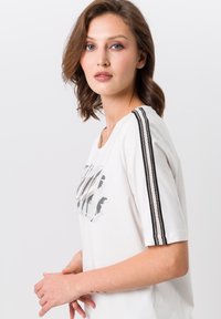 Witte t-shirt met korte mouwen, voorzien van een gestructureerde zwarte opdruk aan de voorkant en twee zwarte strepen langs de schouders. Gemaakt van zacht katoen.