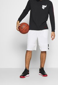 Czarna koszula z długim rękawem z logo byka, w połączeniu z białymi szortami koszykarskimi z czarnym logo Nike oraz czarno-czerwonymi butami sportowymi.