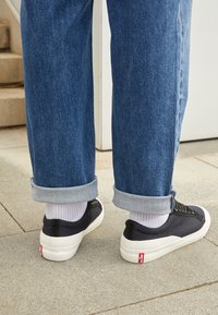 Svarta låga sneakers i canvas med vita gummisulor och svarta skosnören, använda med ljusgråa uppvikta denimbyxor och vita strumpor.