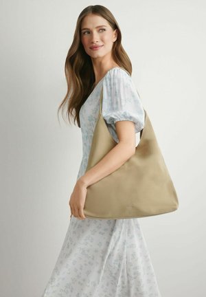 Kobieta o długich brązowych włosach, ubrana w jasnoniebieską sukienkę w kwiaty, niosąca dużą beżową torbę na ramię, spoglądająca lekko w lewo.