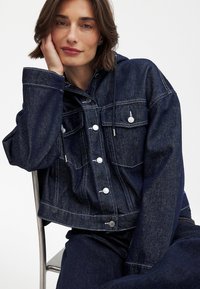 Tumši denima jaka ar kapuci, četri priekšējie kabatas, sudraba pogas un baltas šuves detaļas. Audums rāda gludu, strukturētu izskatu.