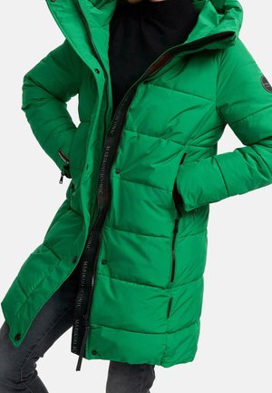 Osoba ubrana w długą zieloną, pikowaną zimową kurtkę z kapturem i czarnym suwakiem, z rękami w kieszeniach, w połączeniu z ciemnymi spodniami.