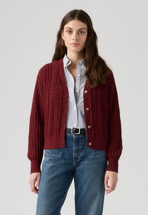 Woman with long dark hair wearing gold hoop earrings, a red knitted cardigan over a striped shirt, blue jeans, and a black belt.