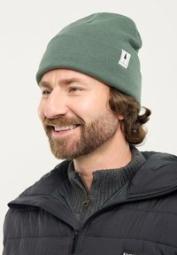 Man with brown beard wearing green knit beanie and dark jacket smiling, facing left on a plain light background.