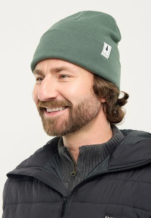 Man with brown beard wearing green knit beanie and dark jacket smiling, facing left on a plain light background.