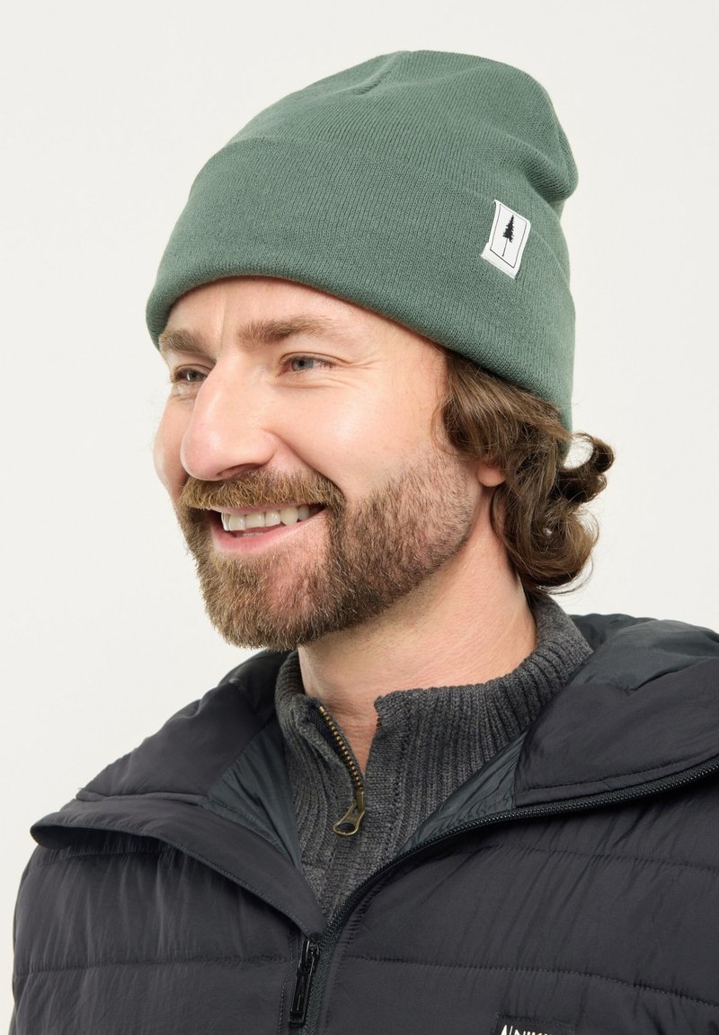 Man with brown beard wearing green knit beanie and dark jacket smiling, facing left on a plain light background.