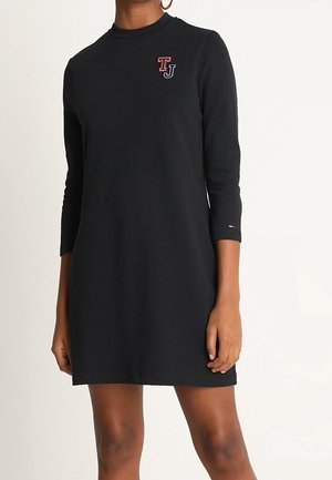 Robe noire à manches longues jusqu'aux genoux avec logo TJ sur la poitrine et petit détail de drapeau sur la manche, portée par une personne debout à la peau brune.