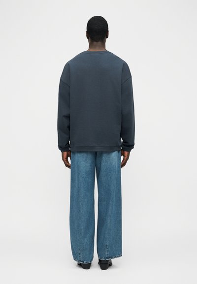 Άτομο με κοντά σκούρα μαλλιά, που φοράει μπλε σκούρο μακρυμάνικο πουκάμισο και φαρδιά μπλε τζιν, στέκεται γυρισμένο πλάτη σε έναν απλό φωτεινό φόντο.