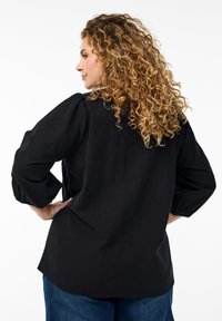 Schwarze Bluse mit langen Ärmeln und Puffärmeln, weicher Textur, abgerundetem Saum und lockerer Passform, kombiniert mit blauen Jeans.