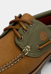 Braune Lederschuhe mit grünen Stoffakzenten, featuring Tan-Schnürsenkel, Metallschnürlöchern und einem aufgenähten Logo an der Seite. Strukturiertes Oberflächendesign.
