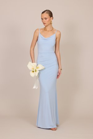 Femeie într-o rochie mulată bleu deschis fără mâneci, ținând crini albi, purtând cercei de tip cerc din aur și colier pe un fundal bej.