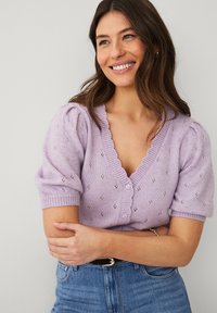 Smilende kvinde med bølget brunt hår iført en lavendelfarvet strikket cardigan med korte ærmer og blå jeans, stående foran en ensfarvet lys væg.