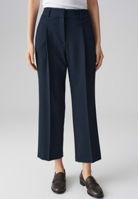 Pantalons larges navy avec une texture lisse, des plis avant et des passants de ceinture, associés à des mocassins marron foncé. Arrière-plan clair et uni.