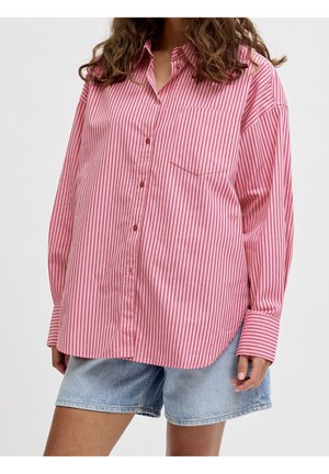 Femme portant une chemise ample à boutons à rayures verticales roses et rouges avec une poche, associée à un short en denim bleu clair.