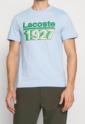 Jasnoniebieska koszulka z zielonym i żółtym napisem "Lacoste 1927" noszona przez osobę z odsłoniętymi ramionami i ciemnymi spodniami.