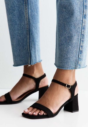 Feet wearing black ankle-strap block-heeled sandals paired with light blue cropped jeans with frayed hems on a white surface.