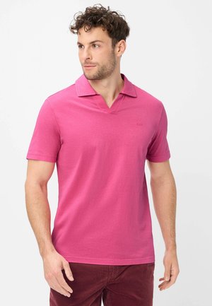 Mann mit lockigem braunem Haar, der ein leuchtend pinkes kurzärmeliges Poloshirt und dunkelrote Hose trägt und nach links blickt.