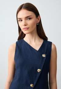 Vestito senza maniche in lino blu navy con design asimmetrico, dotato di bottoni dorati a forma di conchiglia e una finitura liscia e testurizzata.