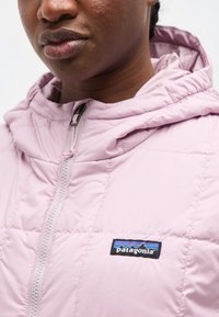 Κοντινό πλάνο ενός ατόμου που φοράει ένα ελαφρώς μοβ καπιτονέ μπουφάν Patagonia με κουκούλα και φερμουάρ μπροστά, με το λογότυπο της μάρκας στο στήθος.