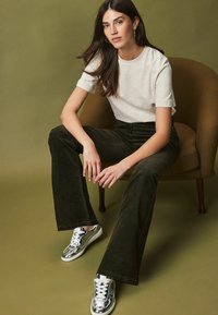 Mörkgröna kordflorsbyxor med vida ben, ljusgrå t-shirt med kort ärm och glänsande silverfärgade sneakers. Modell sitter på en brun stol mot en grön bakgrund.