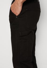 Pantalones cargo negros de corte slim, con múltiples bolsillos, una textura suave y detalles en metal en los botones. La vista lateral muestra la disposición de los bolsillos.