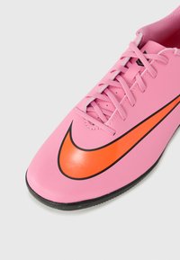 Rozā sintētiska futbola kurpe ar melnu un oranžu Nike swoosh, saistošs dizains un teksturēta zole labākai saķerei.