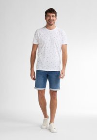 Bílý tričko s malými, barevnými vzory, spárované s vyhrnutými džínovými šortkami. Model má na sobě bílé tenisky. Jednoduchý, ležérní design.