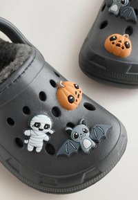 Les sabots en résine grise présentent trois charms décoratifs : une momie blanche, une chauve-souris grise et une citrouille orange, le tout sur un fond texturé.