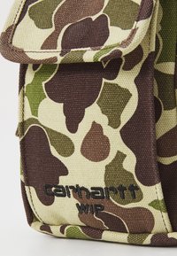 Taška z textilu s maskáčovým vzorom v odtieňoch hnedej, zelenej a béžovej, so zakrývacím uzatváraním. Obsahuje čierne logo "Carhartt WIP". Textúrovaný plátnový materiál.