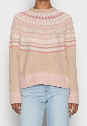 Personne portant un pull en maille beige et rose à motif avec poignets et ourlet côtelés, associé à un jean bleu clair, debout devant un fond uni.