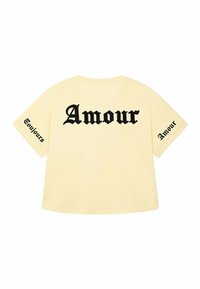 Geel katoenen T-shirt met oversized pasvorm. Tekst op de achterkant in het zwart: "Amour." Aan de linkermouw staat de aanvullende tekst "Toujours." Eenvoudig ontwerp.