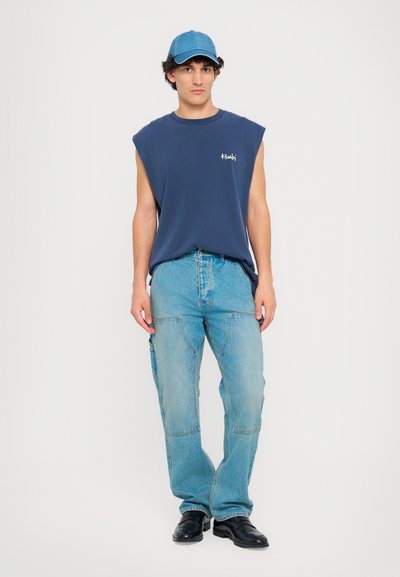 Blå ærmeløs skjorte med logo, parret med lyseblå cargo-jeans. Modellen bærer en blå kasket og sorte sko, sat mod en hvid baggrund.
