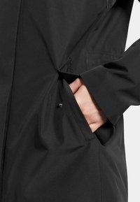 Svart vattentålig jacka med en slät yta. Har en sidoficka och en diskret logotyp. Närbild på hand i fickan.