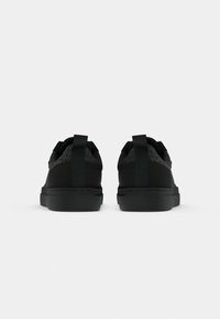 Adidași negri cu un țesut texturat, având un tab posibil la călcâi și o talpă din cauciuc neted. Design simplu cu un finisaj monocromatic.