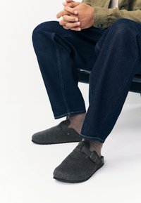 Chaussures en feutre gris à enfiler avec un bout arrondi, une bride ajustable et une semelle extérieure en caoutchouc noir, portées avec un pantalon en denim foncé et des chaussettes marron.