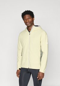 Žlutá lehká bunda s kapucí a zipem, s bočními panely z jemné síťoviny a elastickými manžetami, spárovaná s tmavými upnutými kalhotami.