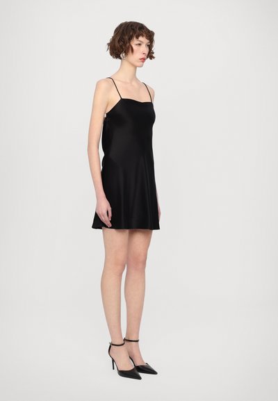 Abito in satin nero con spalline sottili, silhouette svasata e texture liscia. Indossato con tacchi neri a punta e cinturini alla caviglia.