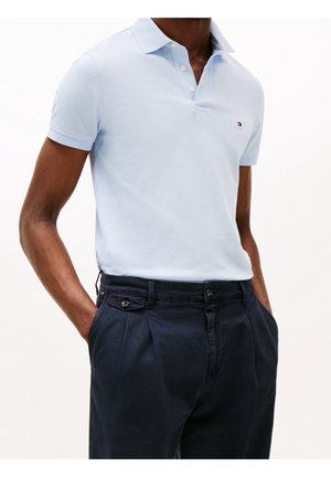 Homme portant un polo bleu clair à manches courtes et un pantalon bleu marine foncé, les mains dans les poches, sur un fond uni.